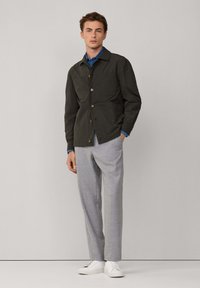 Giovane uomo che indossa una giacca abbottonata verde scuro sopra una camicia blu, pantaloni grigio chiaro e sneakers bianche, in piedi contro uno sfondo semplice.