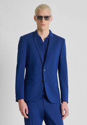 Jonge man met blond haar, die donkere zonnebril en een blauw colbert draagt over een blauw overhemd, tegen een effen lichte achtergrond.