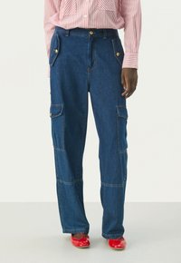 Pantalones cargo de mezclilla azul con un corte relajado, que cuentan con herrajes dorados, bolsillos laterales y costuras en contraste. Llevados con zapatos rojos.