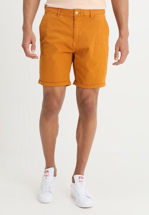Mann trägt orangefarbene Shorts mit hochgekrempeltem Saum und weiße Turnschuhe, steht vor einem einfarbigen hellen Hintergrund.