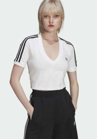 Balta sportinė marškinė su giliu V formos iškirpimu, juoda trimis juostomis akcentais ant rankovių ir mažas juodas logotipas ant krūtinės. Sujungta su juodomis kelnėmis.
