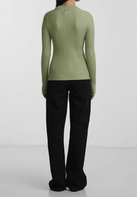 Femme portant un pull à manches longues ajusté et côtelé de couleur vert clair et un pantalon noir ample, debout devant un fond clair uni.