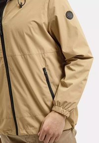 Beżowa lekka kurtka z elastycznymi mankietami, bocznymi kieszeniami na zamek i okrągłym logo z tkaniny na ramieniu. Gładka tekstura, minimalistyczny design.