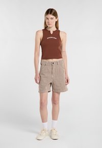 Top sin mangas marrón con logo, pantalones cortos a rayas beige y calcetines blancos. El conjunto presenta un diseño recortado y detalles de botones en los pantalones cortos.