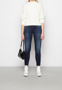 Bílý žebrovaný pletený svetr s širokými rukávy, tmavě modré skinny džíny, bílé tenisky a malá černá kabelka s texturovaným povrchem.