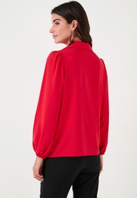 Red blouse with puffed sleeves and a collared neckline. Made from smooth fabric, features a relaxed fit, worn with black pants.