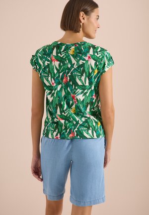Femme portant un haut à manches courtes à motif de feuilles tropicales vertes et un short bleu clair jusqu'aux genoux, debout, dos tourné.