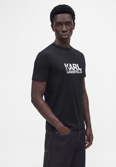Млад мъж, облечен в черна тениска Karl Lagerfeld и тъмни дънки, стои с ръка в джоба пред обикновен бял фон.