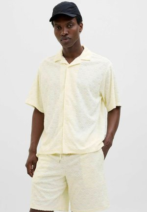 Conjunto de camisa y pantalones cortos a juego en un suave color amarillo claro, con un patrón de olas texturizadas, mangas cortas y un corte relajado. Completa el look con una gorra negra.