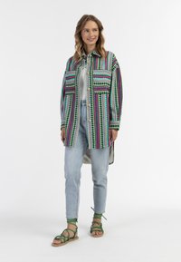 Camicia colorata e stampata in verde, rosa e blu, con una vestibilità rilassata e bottoni. Abbinata a jeans chiari e sandali verde con cinturini.