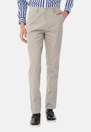 Uomo che indossa pantaloni beige a gamba dritta, scarpe nere e una camicia a righe blu e bianche con un taschino, con una mano nella tasca.