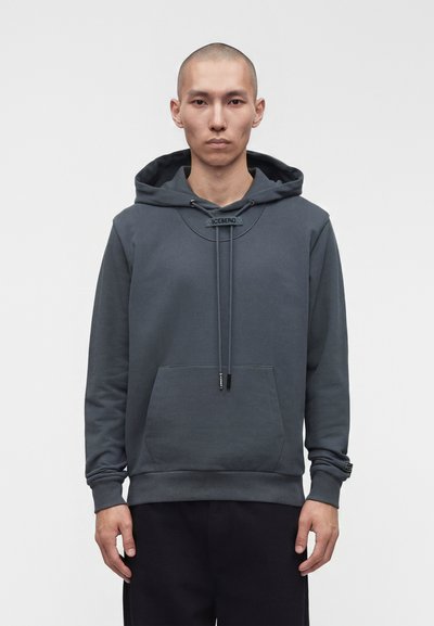 Homme à la tête rasée portant un sweat à capuche gris foncé avec poche avant et cordons, debout devant un fond clair uni.