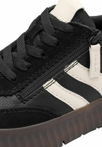 Baskets noires avec des bandes blanches, semelle épaisse texturée, lacets noirs et fermeture éclair latérale avec tirette blanche.
