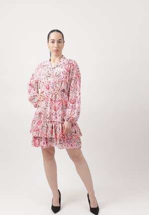 Kobieta stojąca, z jedną ręką na biodrze, ubrana w różową, wzorzystą, wkładaną sukienkę z długim rękawem i falbanami, oraz czarne szpilki, na jednolitym tle.