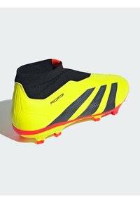 Botín de fútbol amarillo brillante con detalles en franjas negras y un exterior texturizado. Presenta un collar tipo calcetín negro y tacos inferiores rojos.