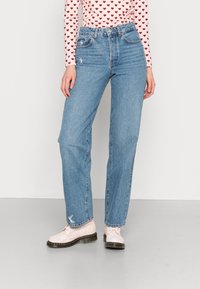 Svijetloplave traperice viskog struka ravnog kroja s izlizanim detaljima, uparene s ružičastim lakiranim cipelama s vezicama.