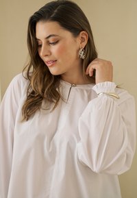 Femme aux longs cheveux bruns portant de grandes boucles d'oreilles argentées, un chemisier blanc à manches bouffantes et un bracelet argenté, regardant vers le bas et souriant doucement.