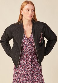 Zwarte bomberjack met ribgebreide manchetten, ritssluiting aan de voorkant en een klein lintdetail. Draagt over een jurk met bloemenpatroon. Glad textuur.