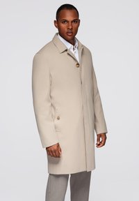 Cappotto impermeabile beige con colletto a faccetta, chiusura a un bottone e tasche laterali, abbinato a pantaloni grigi. Texture liscia.