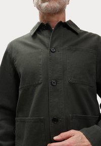 Olive green cotton jacket with a collar, three large black buttons, and two front pockets, featuring a smooth texture and durable construction.