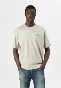 Beige tričko s krátkým rukávem a kulatým výstřihem, na levém prsu s malým modrým logem "BOSS", v kombinaci s vybledlými modrými džíny.