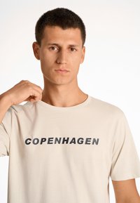 Camiseta beige hecha de tela suave, con cuello redondo y mangas cortas. Texto impreso en negro "COPENHAGUE" exhibido de manera prominente en el frente.