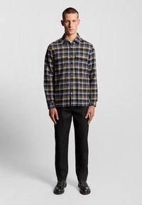 Camicia in flanella a quadri nei colori blu navy, giallo e bianco, con chiusura a bottoni e due tasche sul petto. Abbinata a pantaloni slim-fit neri e stivali.