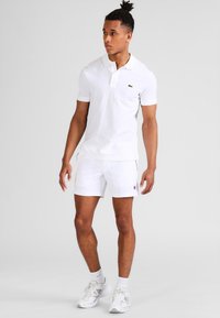 Polo bianco a maniche corte, con un piccolo logo verde; abbinato a pantaloni corti bianchi e sneakers bianche. Tessuto liscio, vestibilità rilassata.