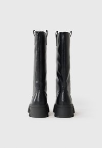 Une paire de bottes noires montantes avec des semelles plates surélevées vues de derrière sur un fond blanc.