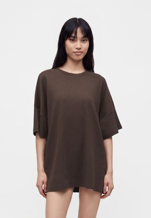 Jeune femme aux longs cheveux noirs portant un t-shirt marron foncé à manches courtes oversize, debout devant un fond blanc uni.