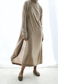 Lungo vestito beige dal taglio rilassato, realizzato in tessuto morbido, con spacchi laterali e scollatura rotonda. Abbinato a semplici scarpe basse marroni.