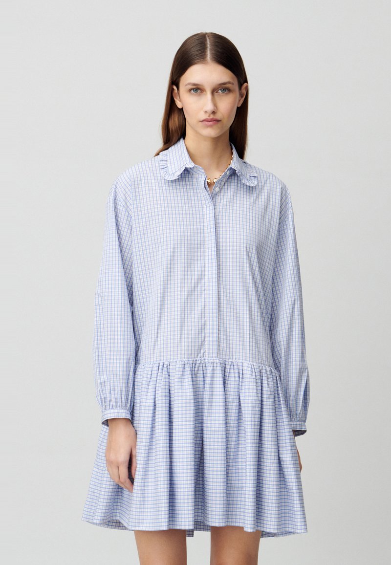 Abito camicia a quadri blu chiaro con maniche lunghe, chiusura frontale con bottoni, orlo arricciato e colletto classico. Tessuto in cotone con texture morbida.