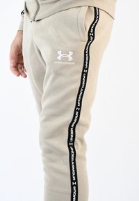 Pantalones de chándal beige de corte slim con un acabado texturizado, franjas laterales negras con la marca "Under Armour" y un pequeño logo blanco en el muslo.
