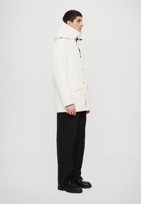 Veste blanche à capuche avec poches avant, boutons noirs et texture lisse, accompagnée de pantalons sombres et de chaussures noires.