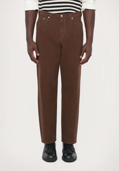 Man in bruin rechte broek, zwarte veterschoenen en een gestreept shirt met opgerolde mouwen, staande tegen een witte achtergrond.