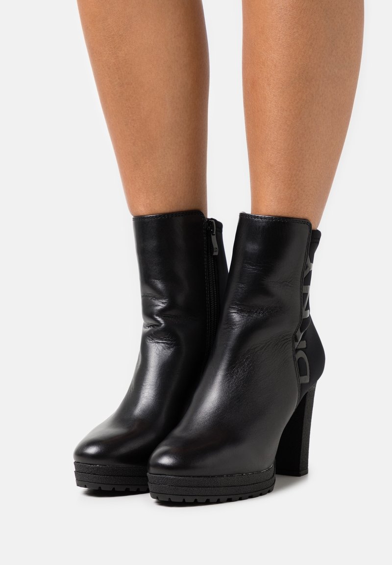 Bottines en cuir noir avec une finition brillante, bout carré, plate-forme texturée et fermeture éclair sur le côté. Présente un logo bien en vue sur le côté.