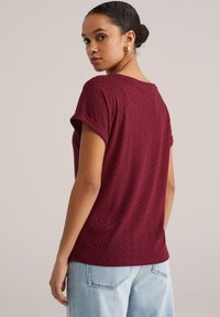 Bordo kratka majica s teksturiranim vzorcem ribje kosti, z zavihanimi rokavi in sproščenim krojem, kombinirana s svetlo modrimi kavbojkami iz jeansa.