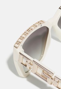 Occhiali da sole bianchi con lenti grigie, caratterizzati da una montatura spessa e un prominente design in catena metallica dorata sulle tempie.