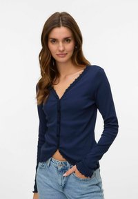 Marinblå långärmad cardigan med V-ringning, spetsdetaljer och knappstängning. Har en figurnära design och scallopdetaljer vid ärmarna och nederkant.