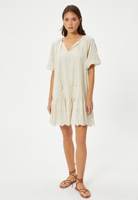 Cremefarbenes Kleid mit V-Ausschnitt, puffigen Ärmeln und ruched, gestuftem Rock. Durchgehendes Häkelmuster. Bänder mit Quasten am Ausschnitt.