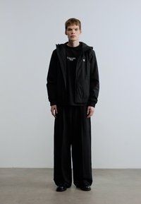 Μαύρο αδιάβροχο μπουφάν πάνω από μαύρο t-shirt, φαρδιά μαύρη παντελόνια και μαύρα παπούτσια. Απλός σχεδιασμός με ελάχιστες λεπτομέρειες, ομαλή υφή.