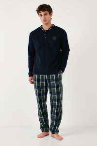 Jaunas vyras basomis stovintis, dėvintis tamsiai mėlyną megztinį su sagomis ir žaliai tamsiai mėlynių languotų pižaminių kelnių, priešais paprastą baltą foną.