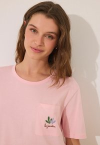 T-shirt di cotone rosa con scollo rotondo, maniche corte e taschino sul petto con piante ricamate in verde e viola e testo.
