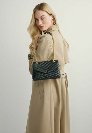 Kobieta o długich blond włosach, ubrana w beżowy trencz, trzymająca czarną pikowaną torebkę na łańcuszku w złotym kolorze, przewieszoną przez ramię.