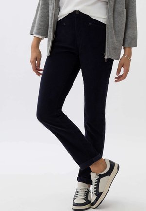 Navy corduroy trousers with a slim fit, featuring a front button closure and rolled cuffs, paired with black and white sneakers.