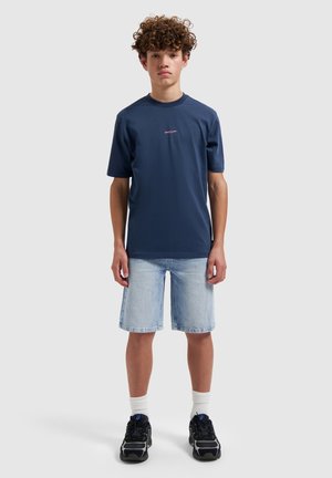 Jonge persoon met krullend haar draagt een marineblauw T-shirt, licht denim korte broek, witte sokken en zwarte sneakers, staand op een effen achtergrond.