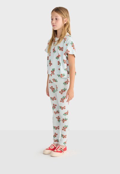 Jeune fille debout portant un haut et un legging assortis blancs avec des motifs de cerises rouges et des chaussures rouges et blanches sur fond blanc.