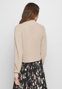 Beiger Strickpullover mit verkürztem Schnitt, gerippten Bündchen und hohem Kragen, kombiniert mit einem schwarzem Rock im Blumenmuster. Weiche Textur sichtbar.