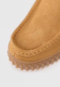 Gros plan sur une chaussure en suède beige avec une semelle en caoutchouc épaisse, présentant un motif de semelle striée et des coutures visibles le long du bord supérieur.
