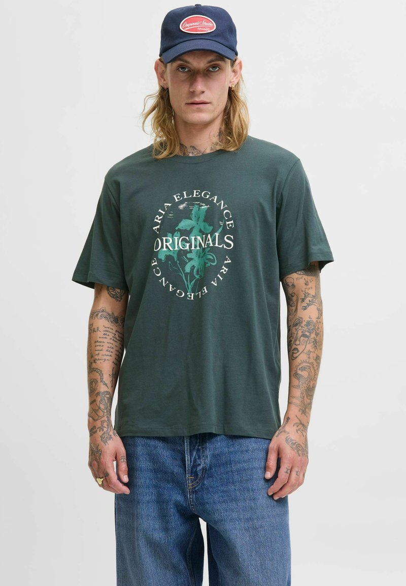 Camiseta de algodón verde oscuro con un gráfico central de diseño floral y el texto "ORIGINALS." Combinada con jeans de mezclilla azul y una gorra azul marino.